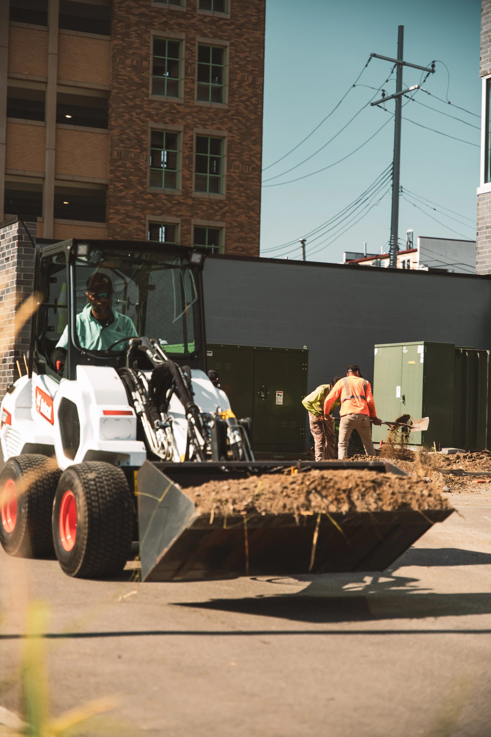 Crew member driving a compact wheel loader with a bucket of soil while two workers shovel dirt beside a commercial building