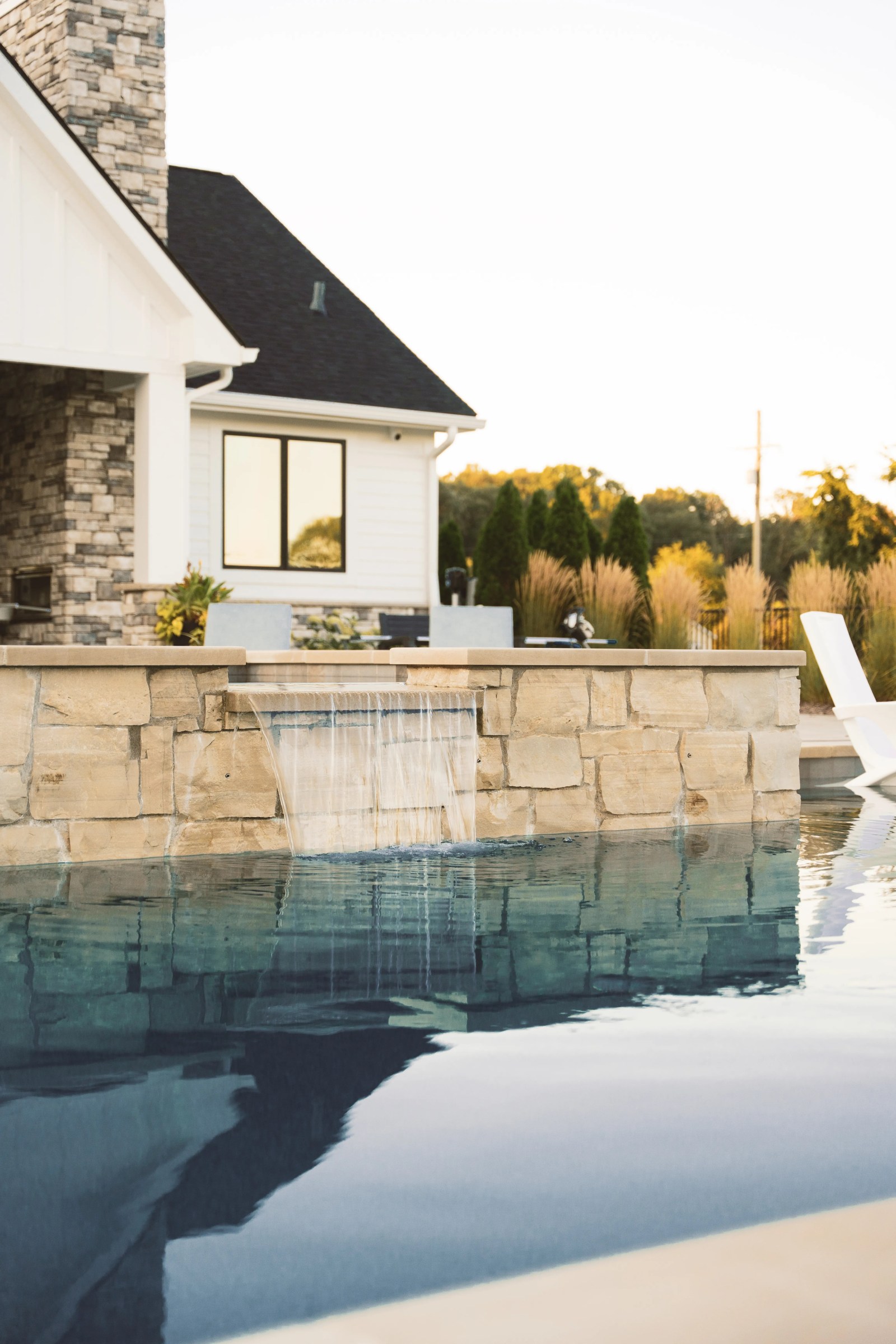 Gunite pool with a stone-faced raised wall and a sheer water feature spilling into the pool beside a white house